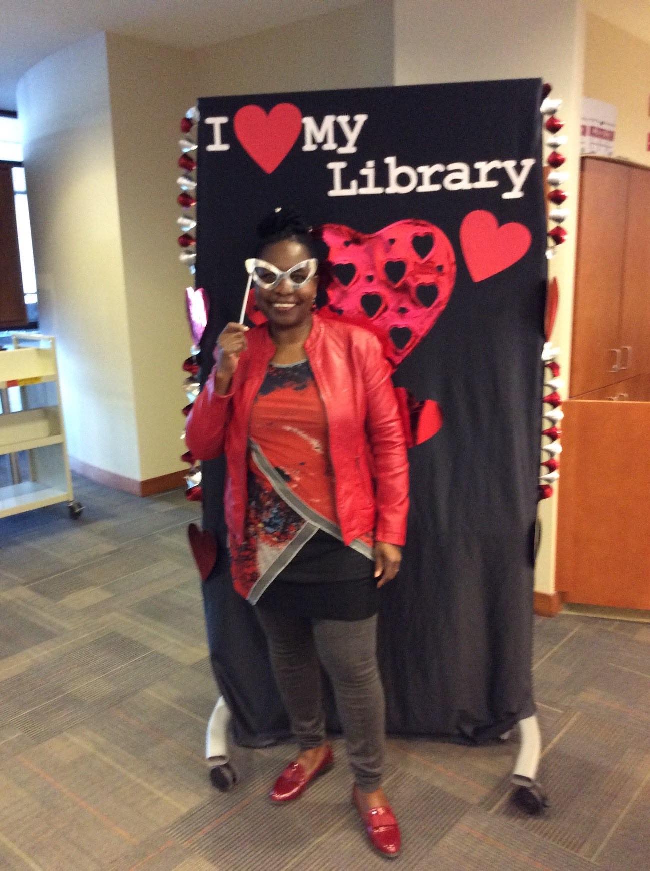 Librarian Mary Chipanshi stands in front of the I love my Library backdrop holding a cut-out of funky glasses up in front of her eyes as though she's wearing them and smiling broadly